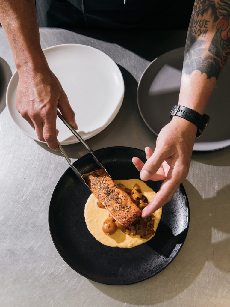 Chef plating seared salmon with tongs over a creamy sauce on a black plate in a commercial kitchen