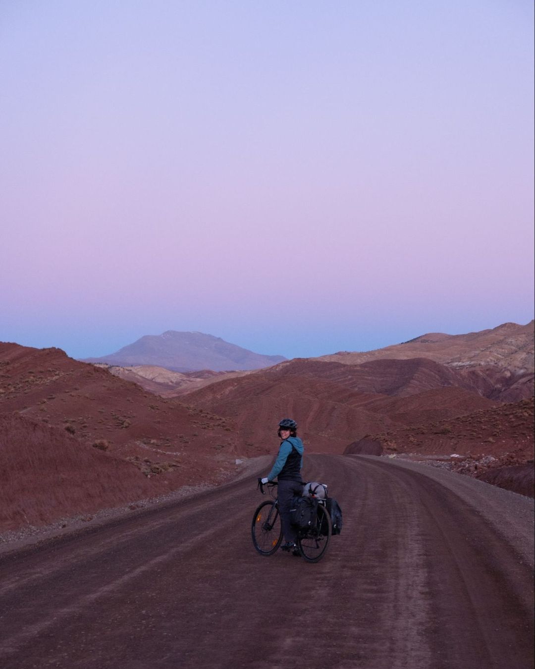 Adult cyclist with helmet pauses on remote dirt road with bike and panniers in desolate, hilly desert landscape under twilight sky.