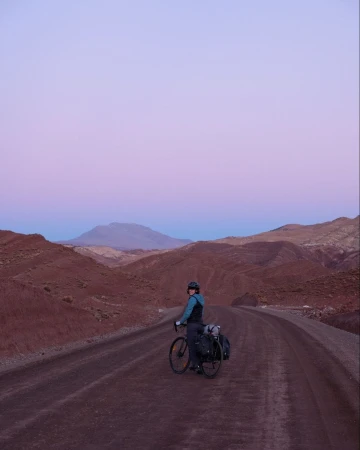 Adult cyclist with helmet pauses on remote dirt road with bike and panniers in desolate, hilly desert landscape under twilight sky.