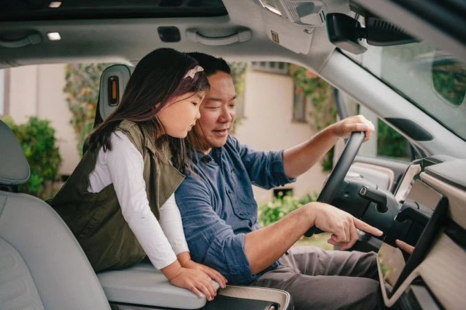 Father and daughter inside a car, the dad pointing at the car's display screen; plants are visible through the car's window.