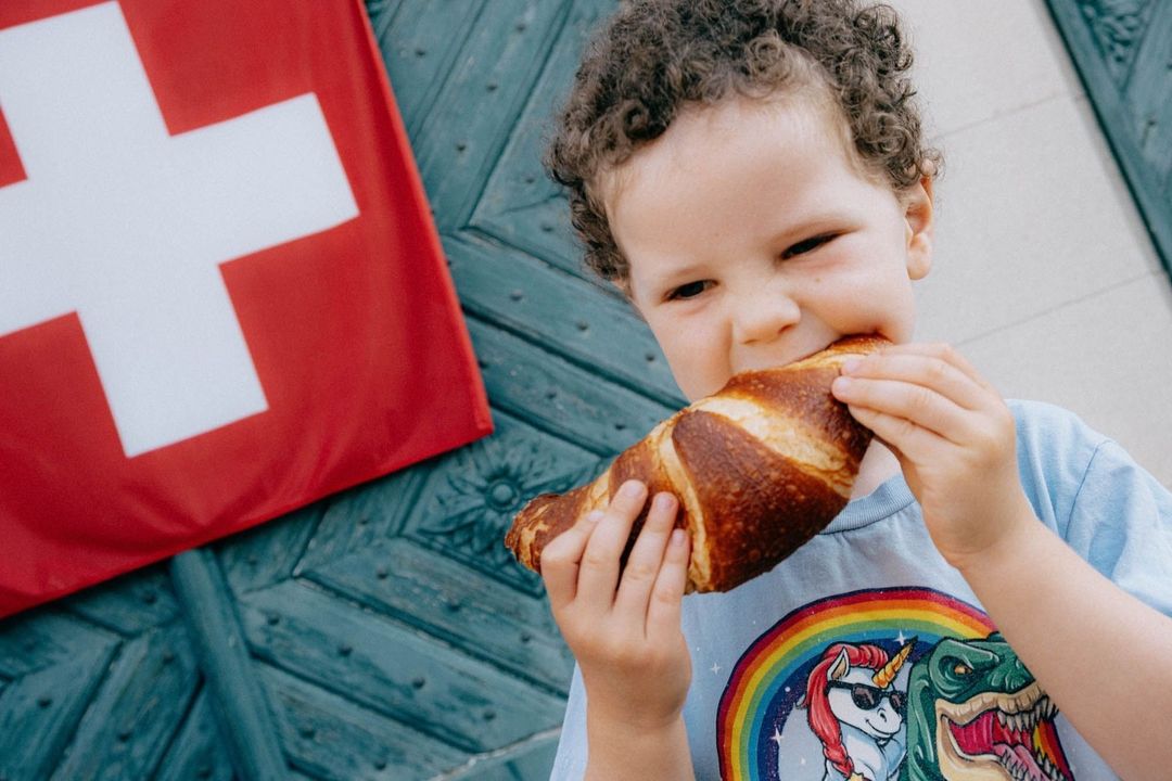 A curly-haired child enthusiastically bites into a pretzel, with a Swiss flag and a blue patterned door in the background.