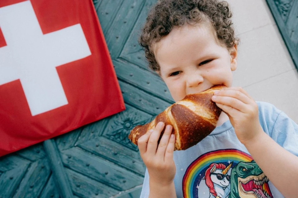 A curly-haired child enthusiastically bites into a pretzel, with a Swiss flag and a blue patterned door in the background.