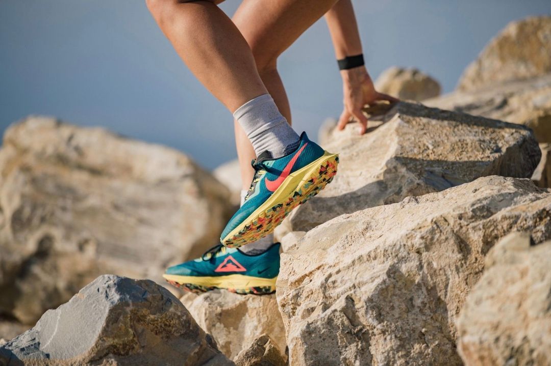 Athletic woman climbs rock formation in trail running shoes; outdoor fitness gear, sport and workout concept. Clear blue sky background.