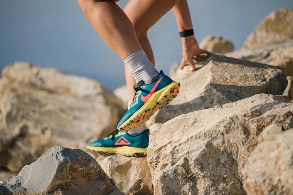 Athletic woman climbs rock formation in trail running shoes; outdoor fitness gear, sport and workout concept. Clear blue sky background.