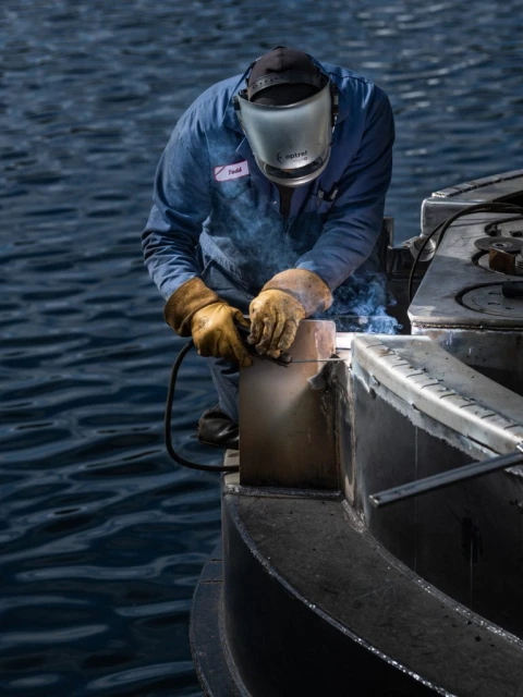 A person wearing protective gear is welding metal on a structure near rippled water, with visible sparks and welding equipment.