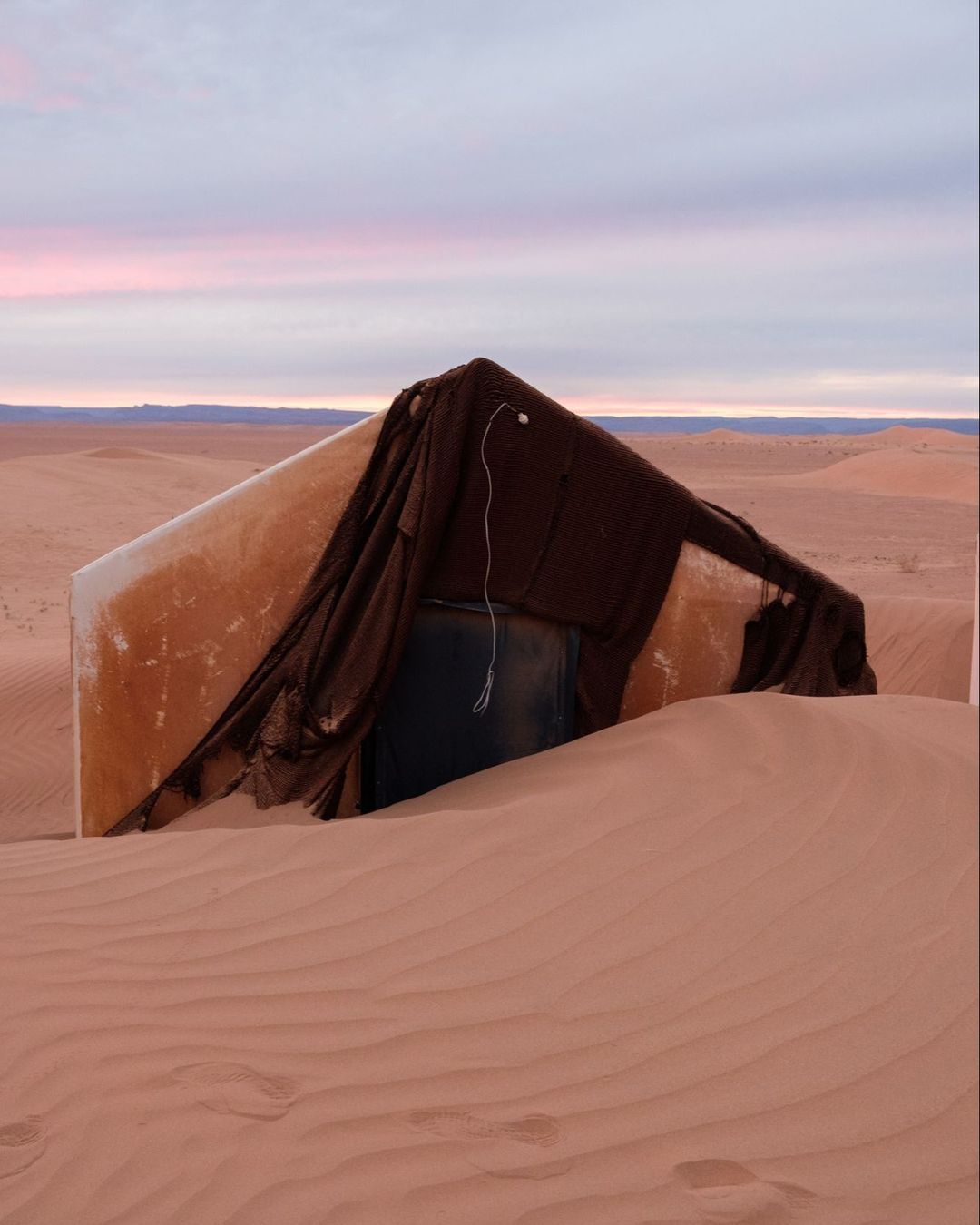 A white structure covered with brown fabric sits on a large expanse of sand dunes under a light pink and purple sky at dusk or dawn.