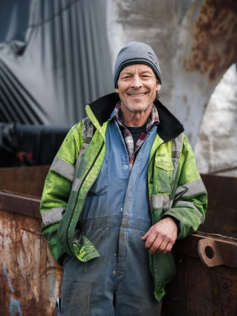 Portrait of a smiling, middle-aged man wearing a beanie, coveralls, and a safety jacket, standing outdoors with industrial equipment in the background.