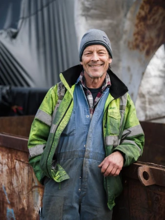 Portrait of a smiling, middle-aged man wearing a beanie, coveralls, and a safety jacket, standing outdoors with industrial equipment in the background.