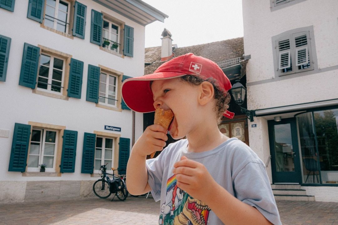 A young child wearing a red baseball cap with a Swiss flag enjoys an ice cream cone outdoors in front of white buildings.