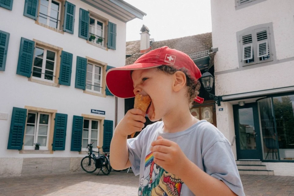 A young child wearing a red baseball cap with a Swiss flag enjoys an ice cream cone outdoors in front of white buildings.