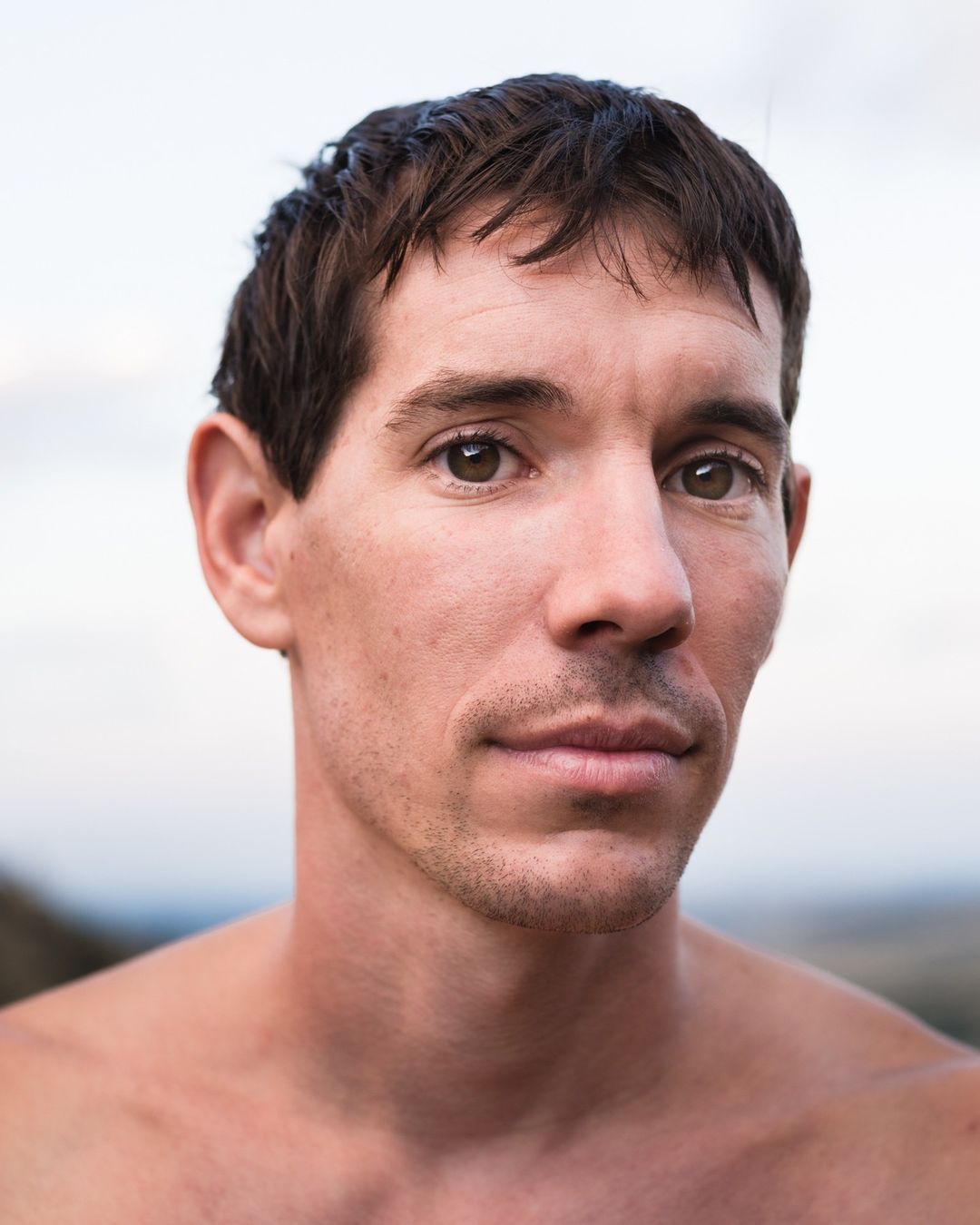 Close-up portrait of a man with dark hair and stubble, looking directly at the camera against an overcast sky background.