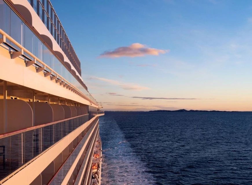 Side view of a cruise ship sailing on calm ocean water under a blue sky with scattered clouds at sunset, promoting luxury travel.
