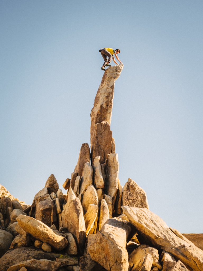 A lone climber summits a tall, thin rock pinnacle, back-lit by a cloudless blue sky. A pile of rocks sits at the base of the pinnacle.