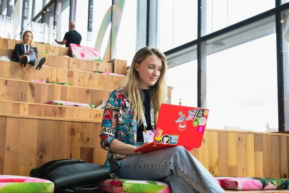 Young woman with blonde hair sits on wooden steps using a red laptop with stickers in a bright open indoor space with other people around.