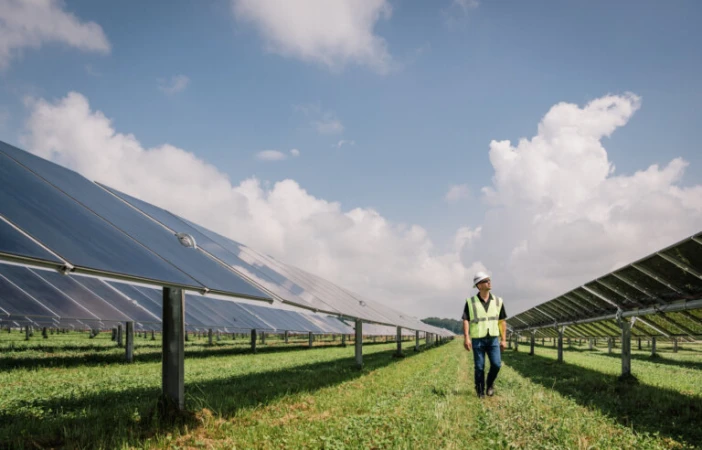 An adult male engineer in safety vest and hardhat walks through a solar panel farm on a partly cloudy day, wide angle, daytime.