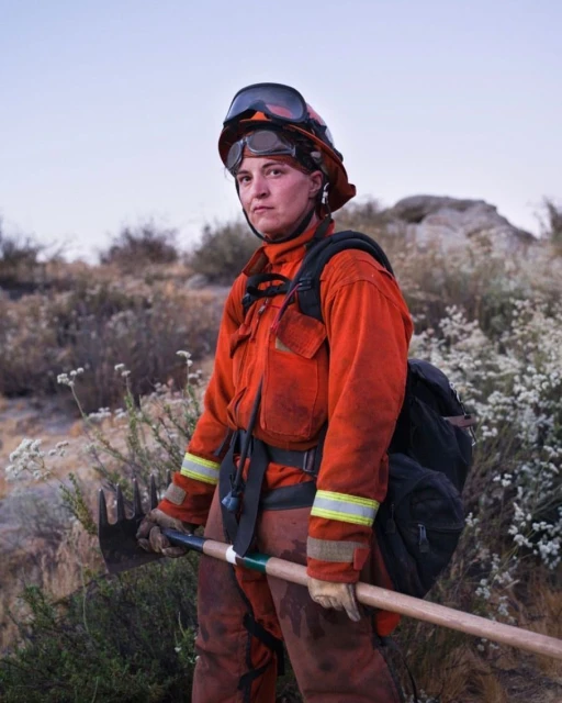 Female firefighter in protective gear stands outdoors on a hillside with a Pulaski axe, under a blue sky, with scattered brush and rocks.