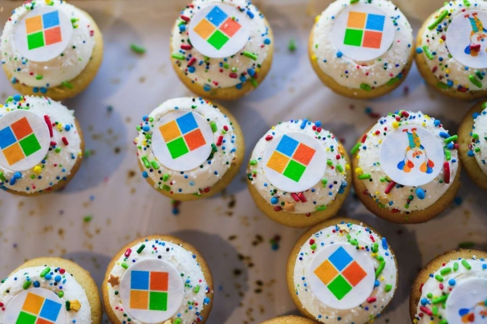 Overhead shot of a batch of frosted cupcakes decorated with sprinkles and logo toppers, arranged on a light-colored surface.