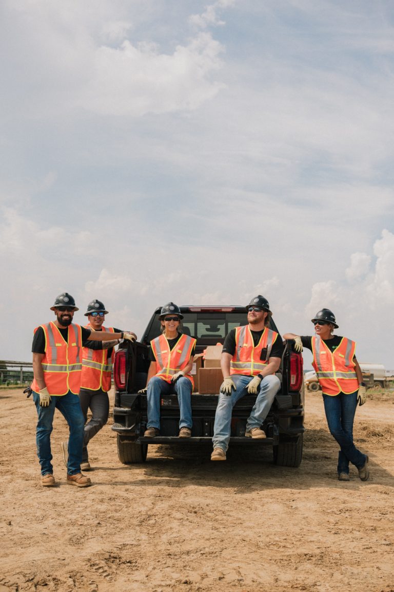 Five construction workers in safety vests and hard hats pose near a pickup truck on a dirt lot under a cloudy sky. Outdoor shot.