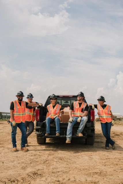 Five construction workers in safety vests and hard hats pose near a pickup truck on a dirt lot under a cloudy sky. Outdoor shot.