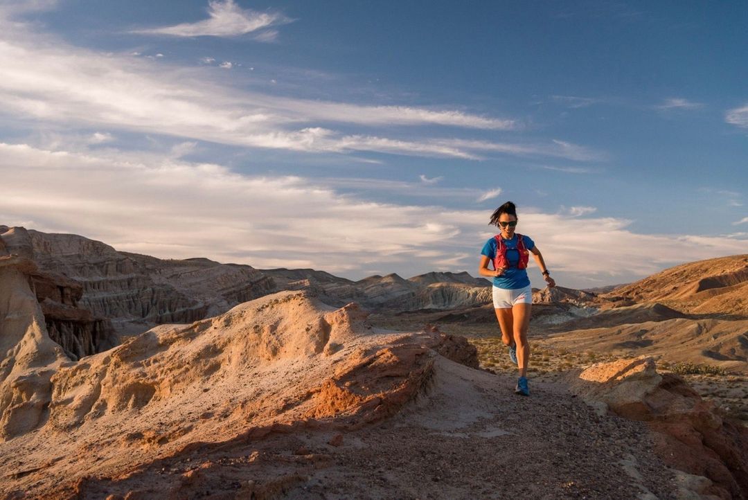 Athletic woman trail running in the desert on a sunny day, wearing a hydration pack, sunglasses, athletic shorts and shirt.