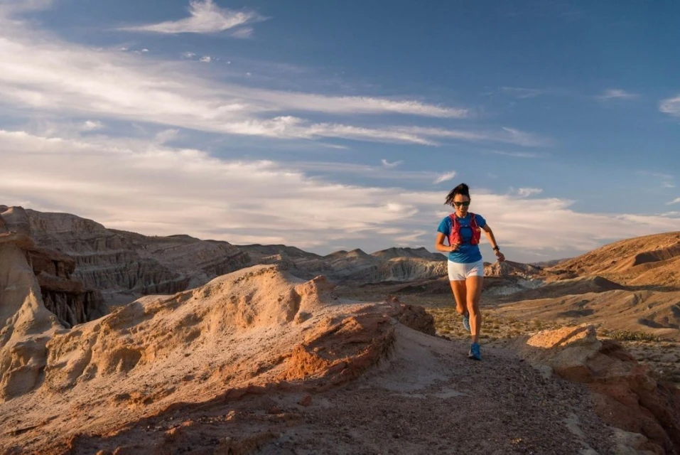 Athletic woman trail running in the desert on a sunny day, wearing a hydration pack, sunglasses, athletic shorts and shirt.