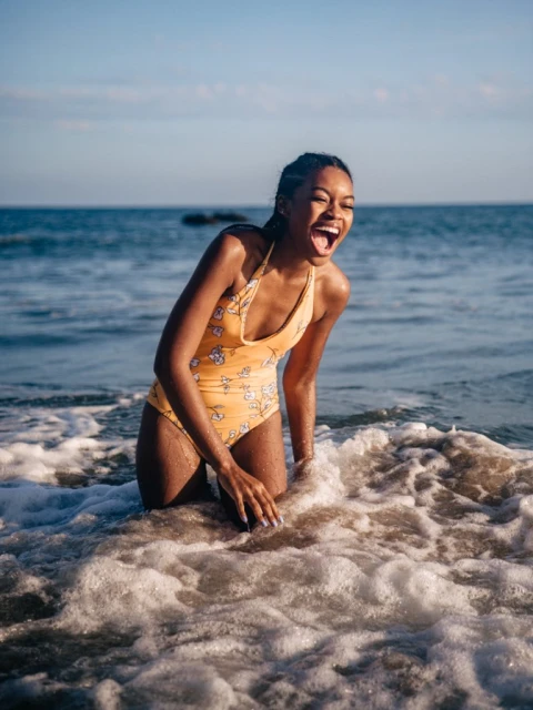 A woman in a yellow swimsuit laughs while standing in the ocean surf. She has dark skin and dark hair, and the background is the sea and sky.