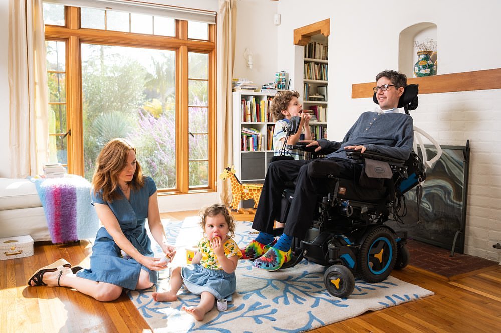 Family portrait featuring a man in a wheelchair, a woman, a young boy, and a toddler sitting on a patterned rug in a sunlit living room.