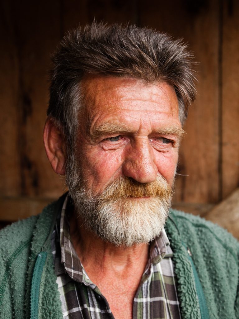 Portrait of a mature adult male with a weathered face, grey beard, and plaid shirt, captured indoors in natural light.