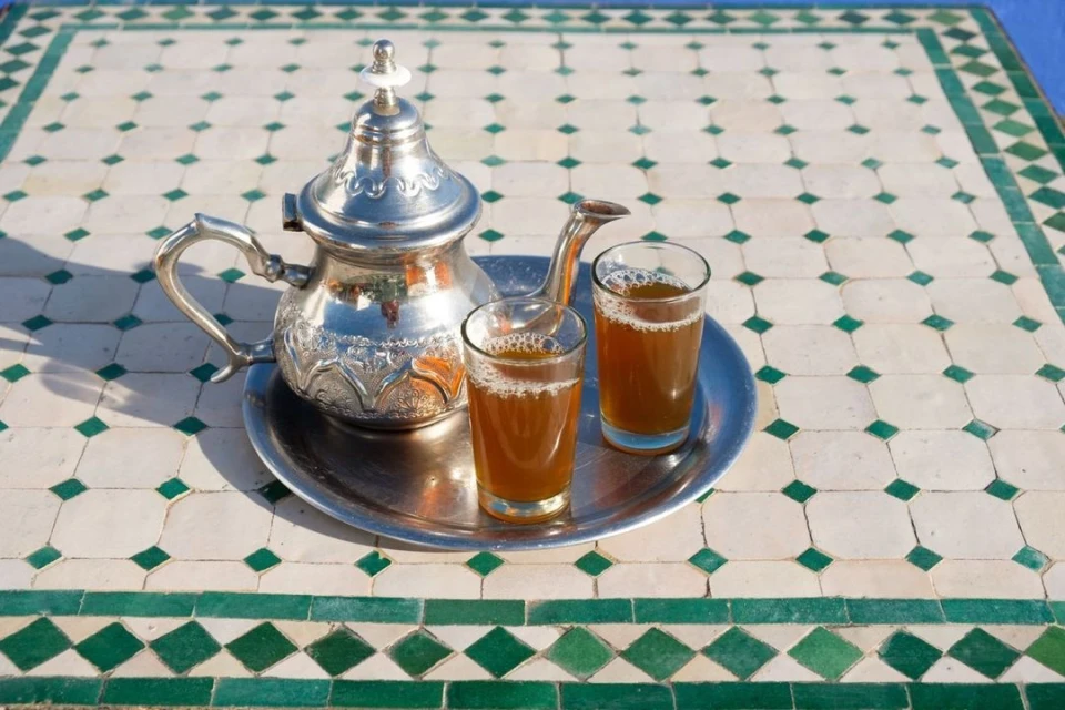 Silver teapot and two glasses of tea on a metal tray, sitting on a tiled table with a white and green geometric pattern.