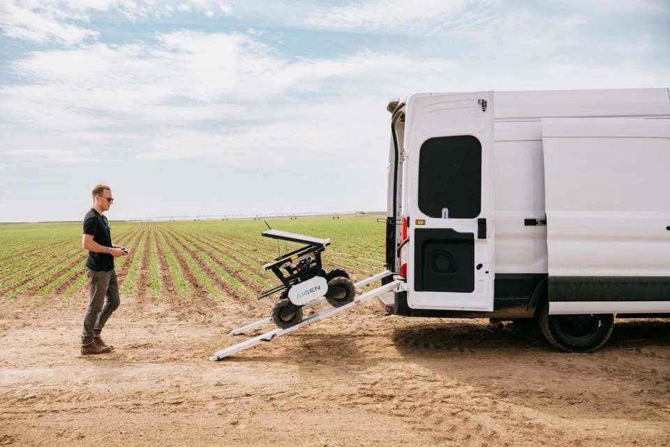 A man operates a robot on a ramp leading to a white van parked in a field, under a partly cloudy sky, showcasing agricultural technology.