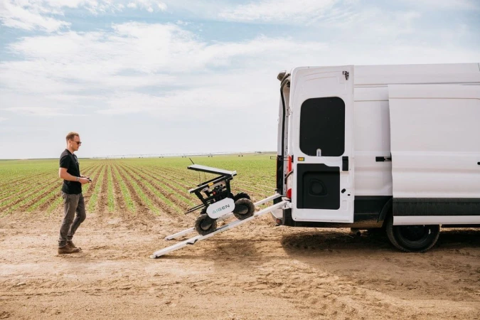 A man operates a robot on a ramp leading to a white van parked in a field, under a partly cloudy sky, showcasing agricultural technology.