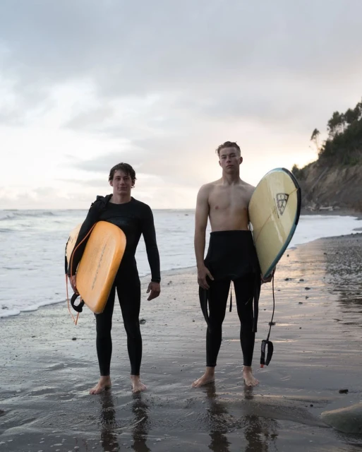 Two young surfers standing on a sandy beach, each holding a surfboard, with a cloudy sky and shoreline in the background.