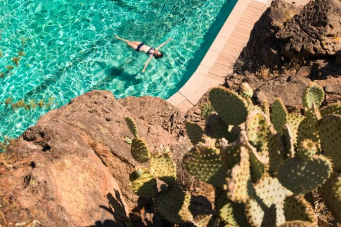High-angle view of a woman floating in a bright turquoise swimming pool bordered by rocky terrain and prickly pear cactuses.