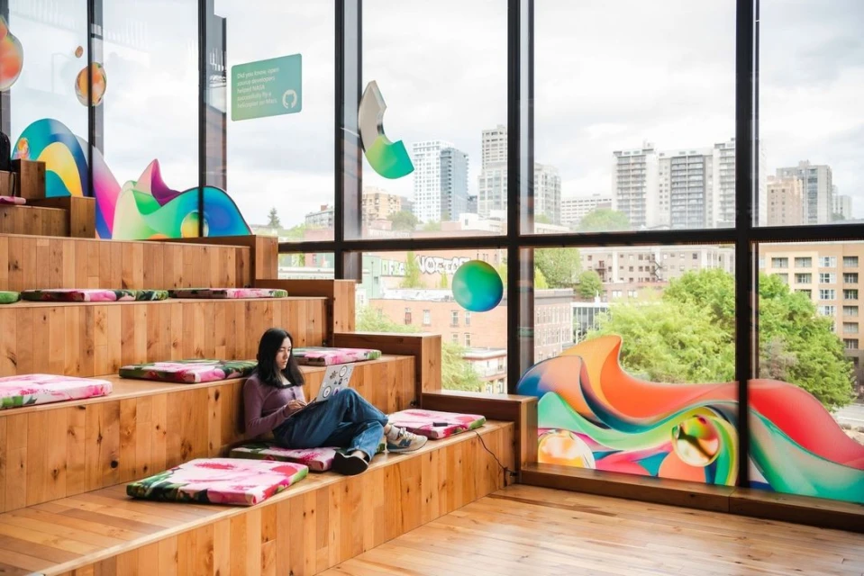 Asian woman sits on a wooden staircase with colorful cushions while using a laptop in a modern office with a city view.