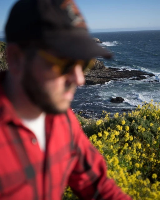 Man in a red plaid shirt and baseball cap stands near yellow wildflowers overlooking the ocean and rocky coastline on a clear day.