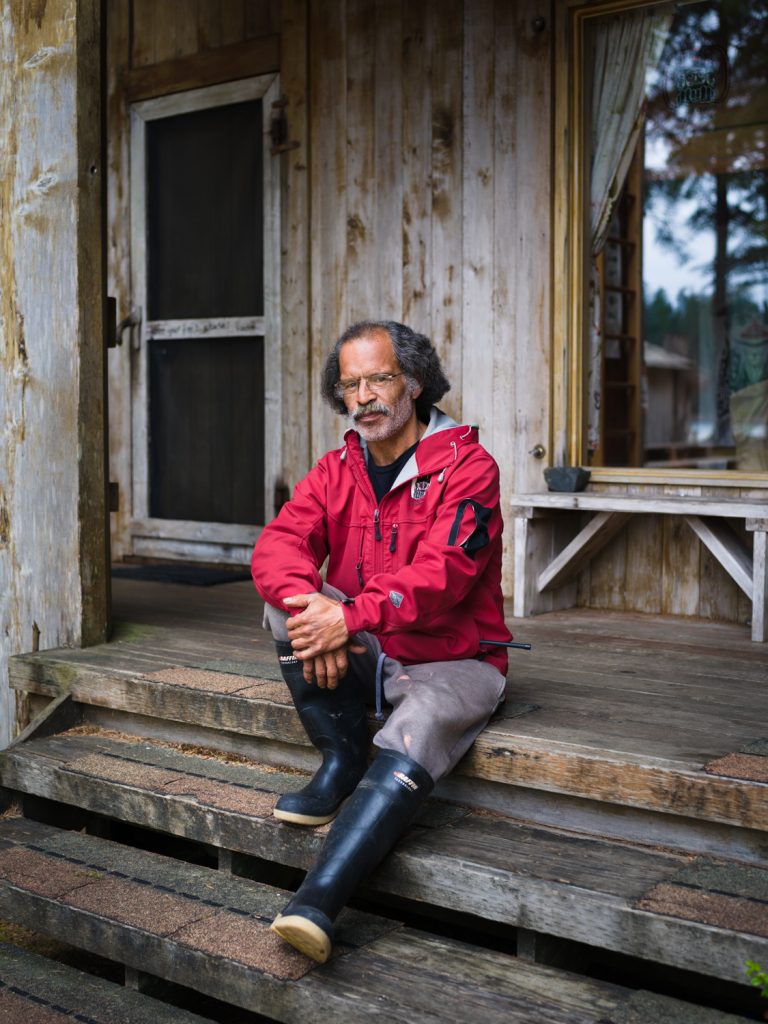 Man with glasses and rubber boots sits on porch steps of a weathered wood cabin in natural light, creating a rustic and relaxed scene.