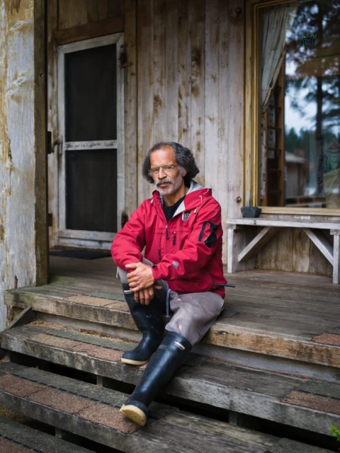 Man with glasses and rubber boots sits on porch steps of a weathered wood cabin in natural light, creating a rustic and relaxed scene.