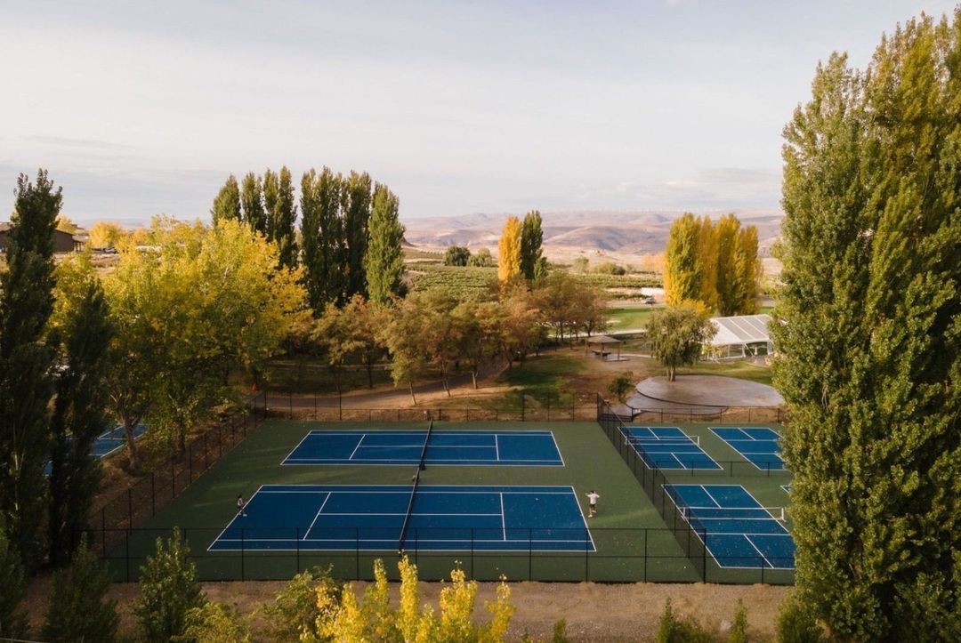 Aerial view of blue tennis and pickleball courts with person playing surrounded by autumn trees and rolling hills on a sunny day.