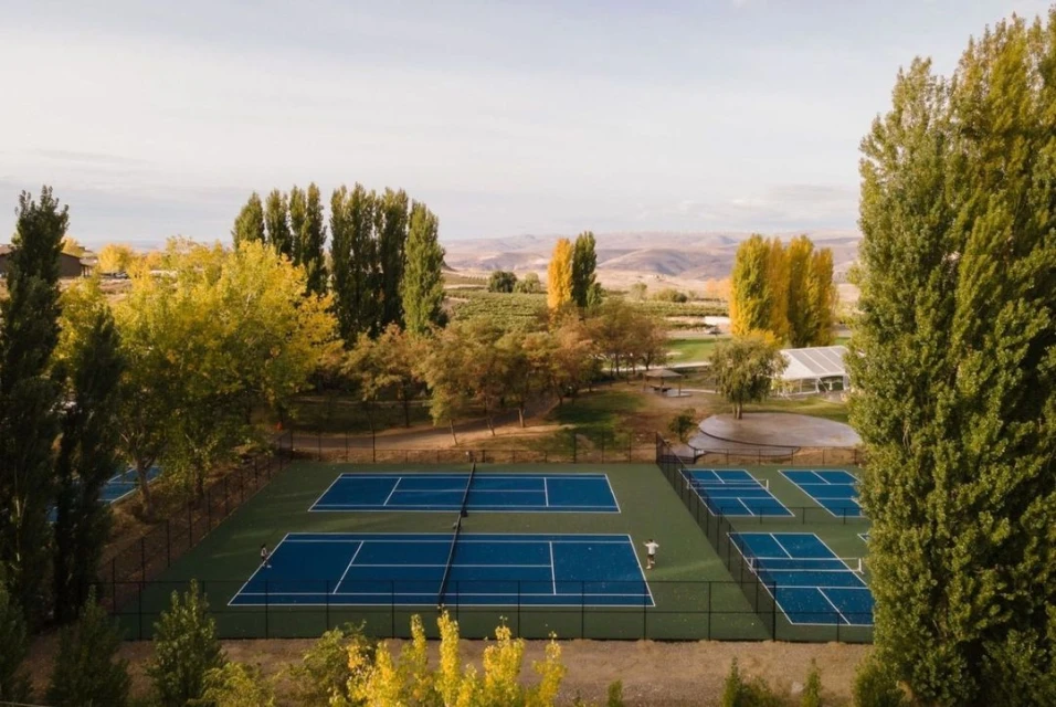 Aerial view of blue tennis and pickleball courts with person playing surrounded by autumn trees and rolling hills on a sunny day.