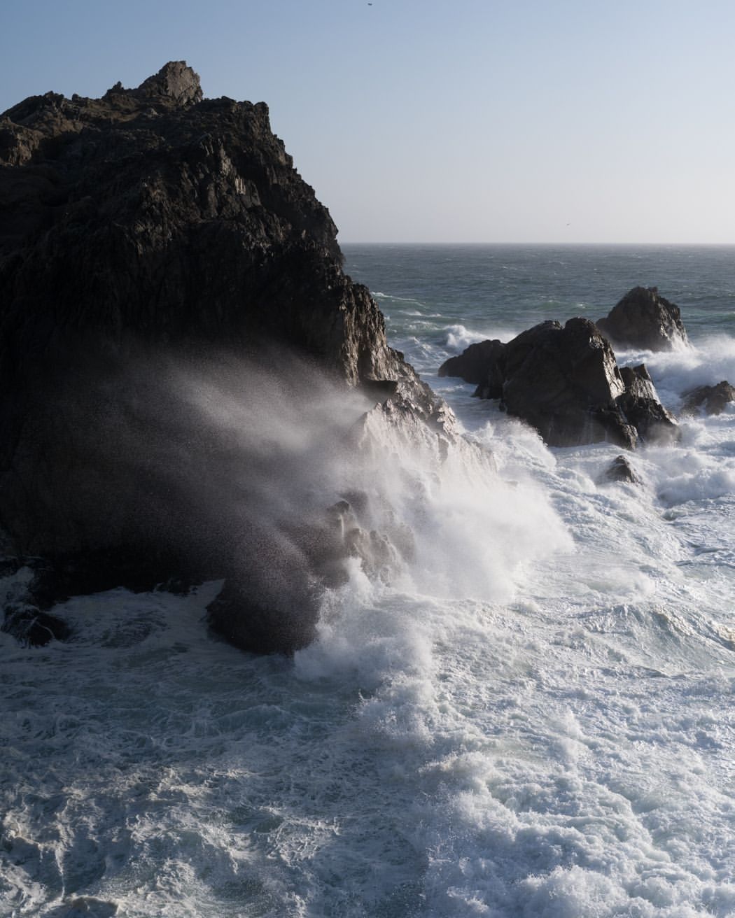 Ocean waves crash against dark rocks on a sunny day. Sea foam and spray fill the air in this seascape.