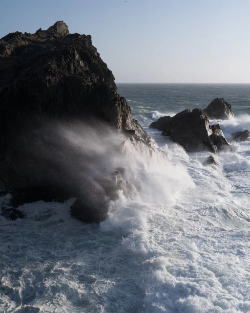 Ocean waves crash against dark rocks on a sunny day. Sea foam and spray fill the air in this seascape.