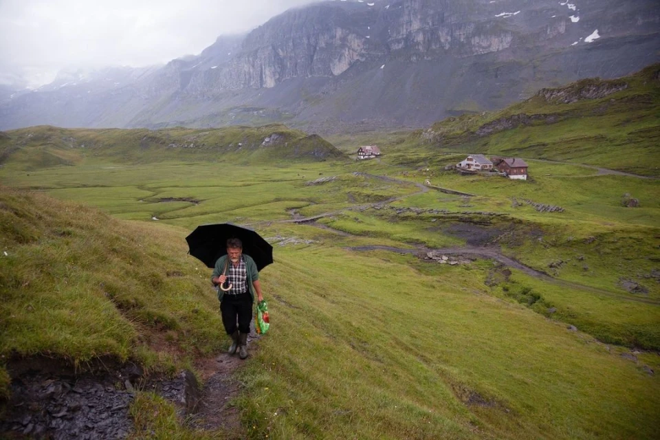 Man walking on grassy hillside path with umbrella in scenic mountain landscape, buildings in distance, overcast sky.