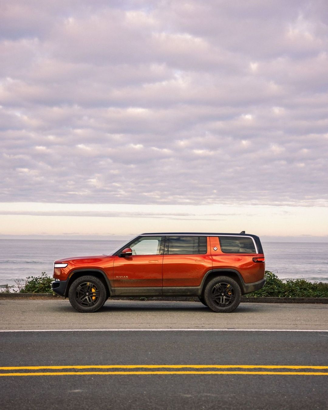 Orange Rivian SUV parked on asphalt road with cloudy sky and ocean in the background, showcasing modern electric vehicle design.