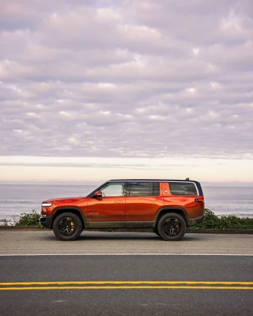 Orange Rivian SUV parked on asphalt road with cloudy sky and ocean in the background, showcasing modern electric vehicle design.
