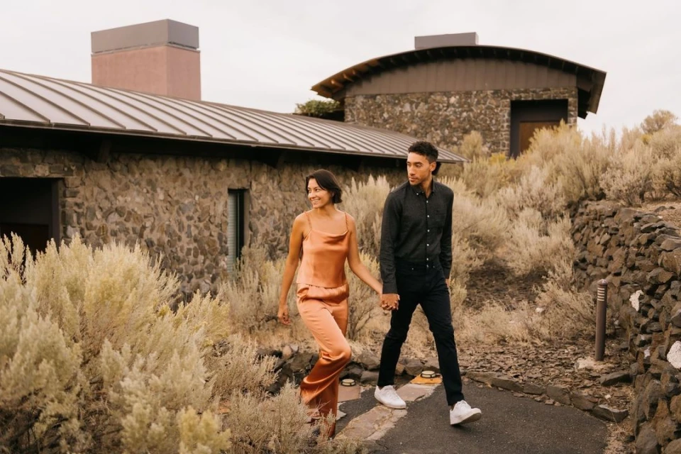 A stylish couple walks hand-in-hand along a stone path, framed by natural foliage and architectural stone buildings.