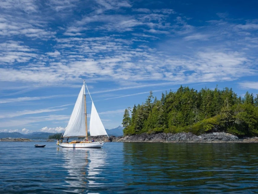 Sailing on tranquil blue waters, a white sailboat glides past a rocky, forested island under a partly cloudy sky, serene maritime scene.