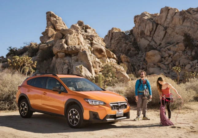 Orange Subaru parked on desert dirt with two rock climbers preparing gear with large rock formations in the background on a sunny day.