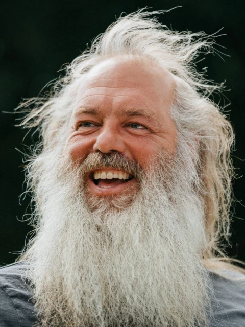 Close-up of a smiling adult male with long white hair and beard, showcasing a joyful expression against a blurred dark background.