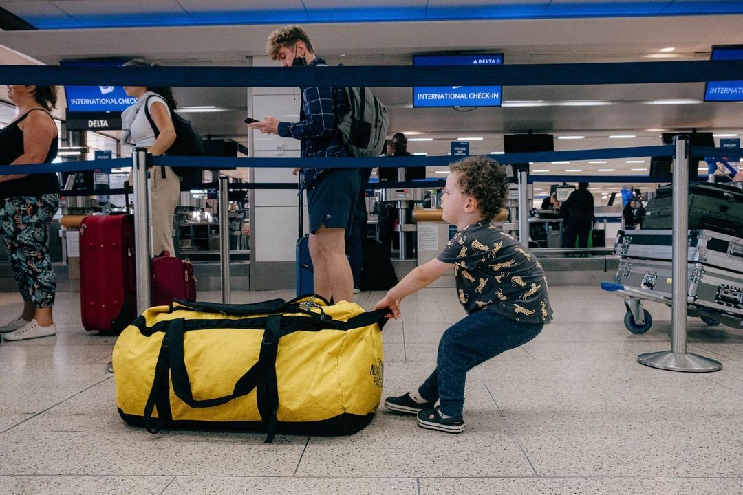 Child pulling yellow duffel bag in airport.  Other travelers in background near check-in.  Interior scene with bright, natural light.
