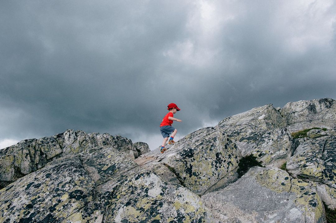 A young child in a red hat and shirt climbs a rocky mountain under a cloudy sky, conveying adventure and the challenges of outdoor exploration.